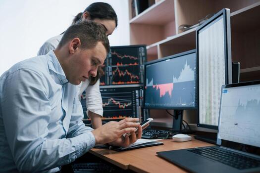 Holdng white smarphone and searching new information. Team of stockbrokers are having a conversation in a office with multiple display screens photo