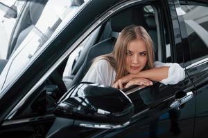 Leaning on the door. Beautiful blonde girl sitting in the new car with modern black interior photo