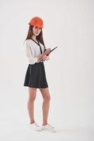 Looking to the camera. Brunette woman in the skirt and orange colored hard hat with notepad in hands stands against white background in the studio photo