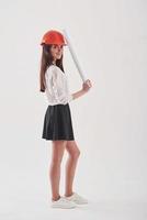 Beautiful legs. Brunette woman in the skirt and orange colored hard hat with paper in hands stands against white background in the studio photo