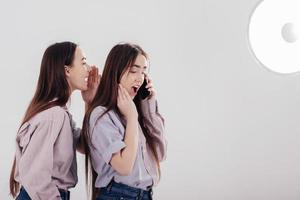 Girl having call and her friend tells what to say. Two sisters twins standing and posing in the studio with white background photo