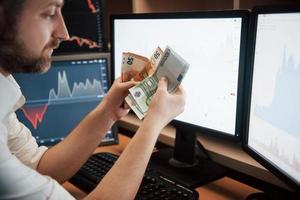Time to get paid. Bearded man in white shirt holds earned money in the office with multiple computer screens in index charts photo