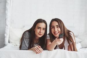 That's moment is so funny, look. Two female twins lying on the white bed at daytime watching movie photo