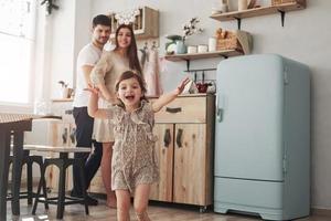 mira, son camarógrafos allí. una niña juguetona se divierte corriendo en la cocina durante el día frente a su madre y su padre foto