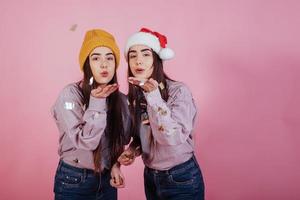 It's Christmas time. Confetti and good mood for all. Air kisses. Two sisters twins standing and posing in the studio with pink background photo