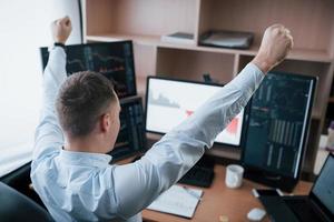 Hands up because of success. Man working online in the office with multiple computer screens in index charts photo
