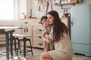 Mother sitting and hugs her daughter at the kitchen at daytime photo