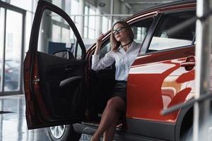 Opening the door. Woman in eyewear, black skirt and white shirt sits in the red automobile photo