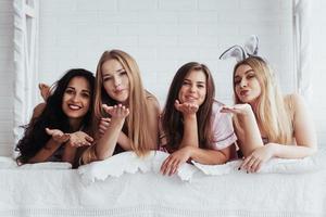 Air kisses. Joyful girls in the nightwear lying on the bed in white room and have celebration photo