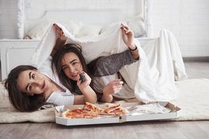 Together is always funnier. Warm blanket. Sisters eating pizza when watching TV while lying on the floor of beautiful bedroom at daytime photo