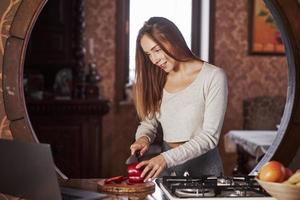 aprendiendo nuevo de internet. mujer joven y guapa parada en la cocina moderna cerca de la estufa de gas y preparando comida usando una laptop foto