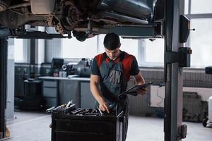 Choosing right instrument. Man at the workshop in uniform fixes broken parts of the modern car photo