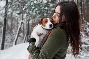 Kissing the pet while holding him on the hands. Smiling brunette having fun while walking with her dog in the winter park photo