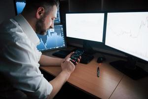 tecnología moderna. hombre barbudo con camisa blanca trabaja usando un teléfono inteligente en la oficina con múltiples pantallas de computadora en gráficos de índice foto