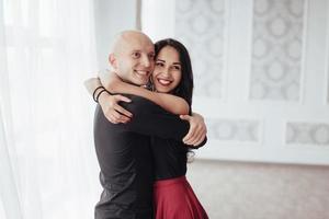 Cute and cheerful people. Hug each other and smiling. Portrait of happy couple indoors. Bald guy and brunette woman stands in the white room photo
