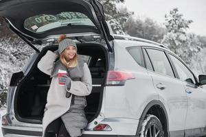 Enjoying the nature. Nice girl sitting at the rear part of modern car in the beautiful winter woods photo