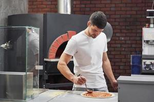 Stove is behind. Baker in white shirt putting sauce to make delicious pizza for an order in restaurant photo