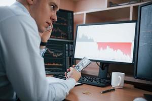 Haves conversation while holding money in the hand. Man working online in the office with multiple computer screens in index charts photo