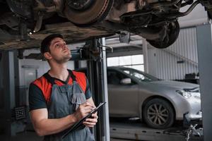 Counting how many details needed. Man at the workshop in uniform using notepad for his job for fixing broken car photo