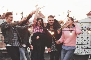 Wind will not prevent us. Playing with sparklers on the rooftop. Group of young beautiful friends photo