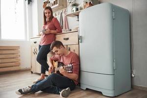 Guy sits on the floor. Young guitarist playing love song for his girlfriend in the kitchen photo