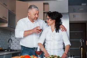 Let's drink some wine. Senior man and his wife in white shirt have romantic dinner on the kitchen photo