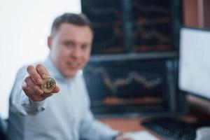 Focused photo. Businessman holding bitcoin in hand while sitting in modern office with many monitors with graphs photo
