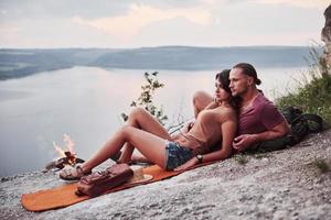 Hugging couple with backpack sitting near the fire on top of mountain enjoying view coast a river or lake. Traveling along mountains and coast, freedom and active lifestyle concept photo