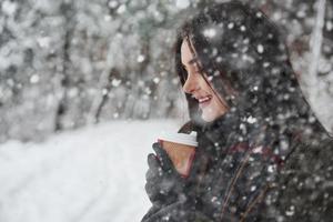 nieve cayendo en el bosque. chica con ropa de abrigo con una taza de café da un paseo por el bosque de invierno foto