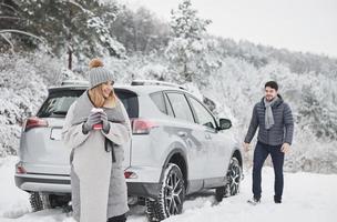 Cheerful couple. Girl is waiting for her husband near their automobile and holding cup with warm drink photo