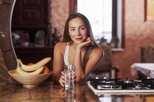 Holding glass with water. Young woman standing in the modern kitchen near gas stove photo