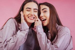 Playing like kids again. Two sisters twins standing and posing in the studio with pink background photo