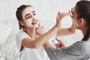 You look funny. Conception of skin care by using white mask on the face. Two female sisters have weekend at bedroom photo