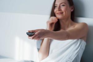 Focus on the remote controller. Pretty young woman sitting on the bed at morning time in her room photo