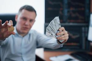 Conception of crypto currency. Businessman holding bitcoin and money in hands while sitting in modern office with many monitors with graphs photo