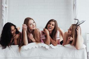 buenos viejos amigos. chicas alegres en ropa de dormir acostadas en la cama en la habitación blanca y celebrar foto