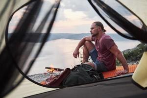 View from tent of traveller with backpack sitting on top of mountain enjoying view coast a river or lake. Traveling along mountains and coast, freedom and active lifestyle concept photo