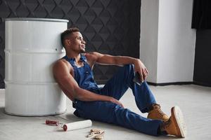 Holding black colored smartphone. Time for the rest. African american man sits on the floor and leaning to the white barrel photo