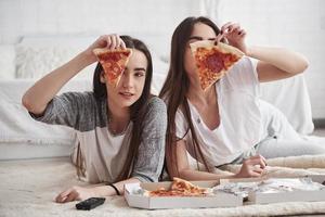 Holds food in the hands. Sisters eating pizza when watching TV while lying on the floor of beautiful bedroom at daytime photo