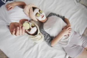 secreto de la belleza de la mujer. acostado en la cama blanca. vista superior. concepción del cuidado de la piel usando mascarilla blanca y pepinos en la cara foto