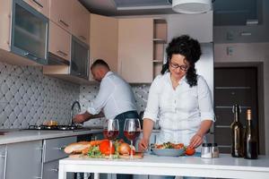 Cute couple. Man and his wife in white shirt preparing food on the kitchen using vegetables photo