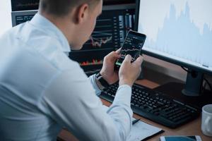Using smartphone. Man working online in the office with multiple computer screens in index charts photo
