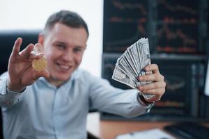 Happy rich man. Businessman holding bitcoin and money in hands while sitting in modern office with many monitors with graphs photo