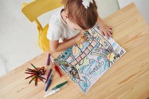 Sitting on the yellow chair. Cute little girl in art school draws her first paintings by pencils and markers photo