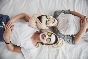 todo el mundo está sonriendo. acostado en la cama blanca. vista superior. concepción del cuidado de la piel usando mascarilla blanca y pepinos en la cara foto