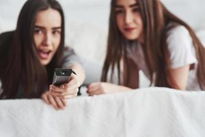 TV remote controller in hands of cheerful girl. Two female twins lying on the white bed at daytime watching movie photo