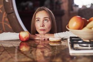 Making funny face. Cute young housewife leaning on the table with apple and cookie and looks up photo