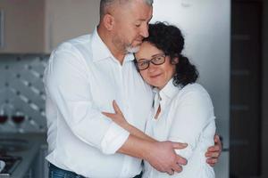 Hugs each other. Senior man and his wife in white shirt have romantic dinner on the kitchen photo