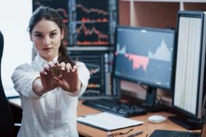 Modern currency. Beautiful woman holding bitcoin in hands while stands in modern office with many monitors with graphs photo