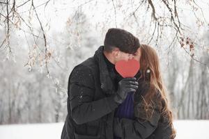 Kiss cencored by red heart. Gorgeous young couple have good time together in snowy forest photo